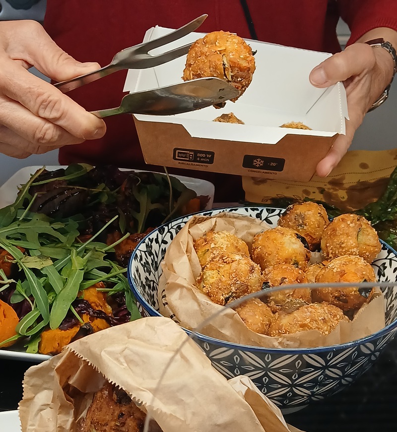 Pinze ripongono polpette impanate in vaschetta da asporto con scritte 800 W e -20°, accanto a ciotola blu e rucola