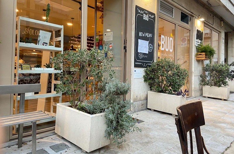 Ingresso di Buò con vetrine, lavagna «New menu», fioriere bianche con ulivo e lavanda, panca in metallo e sedia in legno