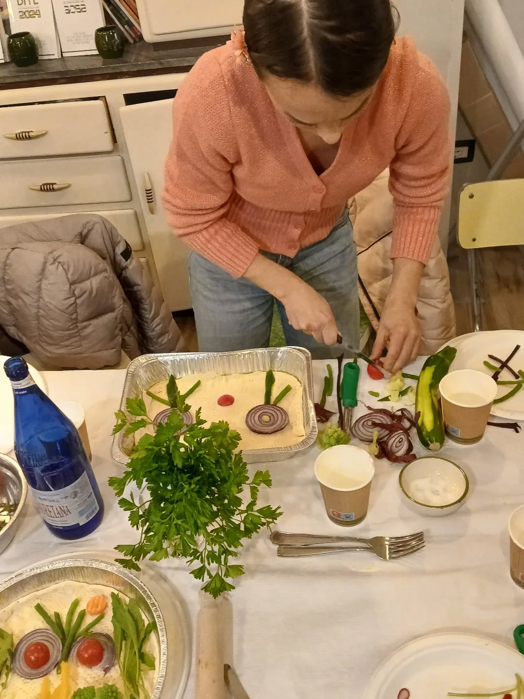 Donna in cardigan rosa taglia verdure su un tavolo con teglie di focaccia decorate a fiori di cipolla e prezzemolo