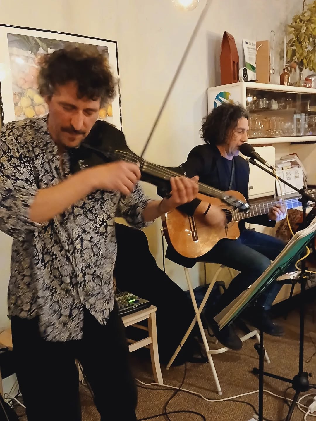 Violinista in camicia a fiori e chitarrista seduto al microfono suonano dal vivo tra mensole con bicchieri e un quadro di limoni